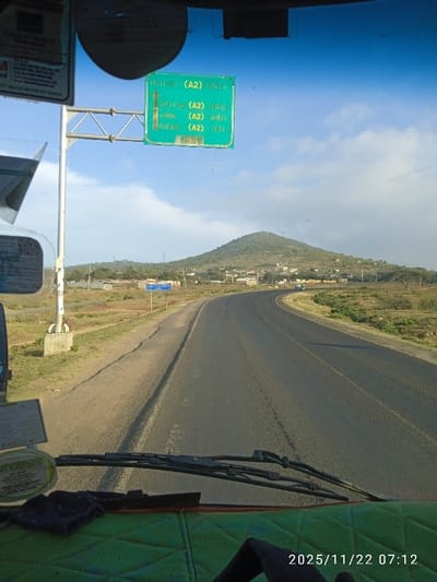 A view from inside a vehicle shows a winding road heading toward a hill. Road signs indicate Nairobi (A2) is 185 kilometers away, with closer distances for other destinations. The vehicle's dashboard is visible, and the date "2025/11/22" with the time "07:12" is displayed at the bottom. The landscape on either side of the road appears dry and grassy, with scattered buildings near the base of the hill.