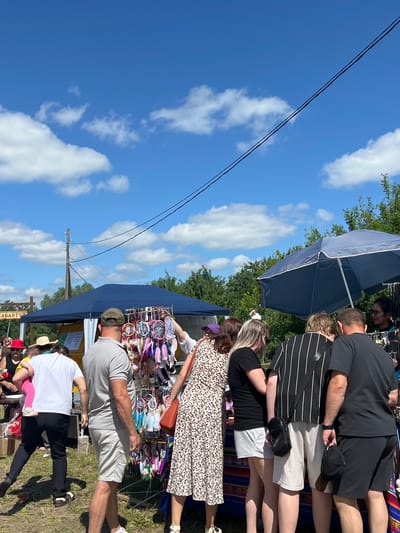 A group of people are gathered around a market stall displaying dreamcatchers on a sunny day in Kalinino, Russia. A blue tent is set up behind the stall, and trees form a backdrop against a bright blue sky with scattered white clouds. Several individuals are examining the merchandise, with one person holding up a dreamcatcher. The ground is covered in dry grass and dirt. Electrical wires are visible stretching across the sky, connected to a wooden utility pole.