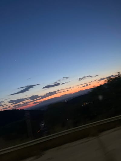 The image depicts a twilight scene in Amygdaleonas, Greece. The sky transitions from deep blue to orange hues near the horizon, with scattered clouds. The silhouette of a dark landmass is visible along the horizon, dotted with faint lights that likely indicate a settlement. A section of a structure, possibly a building or window frame, is seen on the right side.