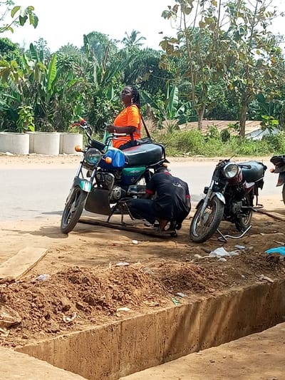 The image depicts an outdoor roadside scene in Aramoko-Ekiti, Nigeria, during daylight hours. In the foreground, a dark-complexioned man in dark work attire crouches on the ground, actively working on the engine area of a blue and dark green motorcycle. The motorcycle is propped on a stand, supported by a wooden plank beneath. Seated on the rear of this motorcycle is a dark-complexioned woman with braided hair, wearing an orange short-sleeved top and large hoop earrings; she holds a small object and looks towards the upper left. A second maroon-colored motorcycle is parked on its stand to the right of the first, unattended. The ground surface consists of reddish-brown dirt with scattered litter, including white paper and plastic, alongside a low, rough concrete barrier. A paved road runs behind the motorcycles. The background features dense tropical vegetation, including banana plants and various trees, beneath a clear sky. Two cylindrical concrete planters are visible on the far left, near the road's edge.