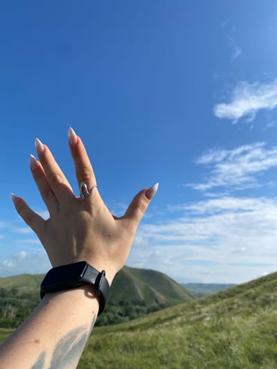 In a close-up shot, a person's left hand is extended towards the sky, with fingers splayed and pointing upwards. The hand has long, almond-shaped nails with white tips and a silver ring with a wavy design on the middle finger. A black fitness tracker is visible on the wrist. The background features a vast, clear blue sky with scattered white clouds, and rolling green hills in the distance. The scene is set in a natural environment, possibly a rural area or a park. There is no identifiable information about the specific location in Andreevka, Russia, or any interactions or actions beyond the static pose.