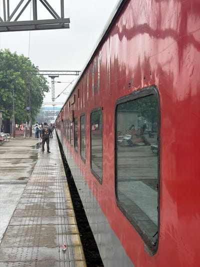 The image depicts a red and grey train at a train station platform in Ballabgarh, India. The platform is wet, with visible puddles and water stains on the concrete tiles, suggesting recent rain. Several people are visible on the platform. In the foreground, a man wearing a dark t-shirt and pants stands facing away from the camera, looking towards the train. Further down the platform, a group of people are congregated, some appear to be waiting or disembarking. Trees line the left side of the platform, and overhead electrical lines for the train are visible. The windows of the train reflect the surrounding environment, showing glimpses of structures and foliage. A discarded red and white disposable cup lies on the platform near the tracks. The sky is overcast.