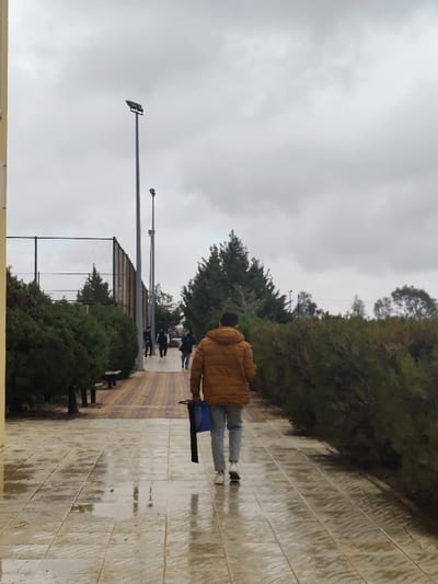 The image depicts an outdoor paved pathway in Jibb as-Safa, Syria, under an overcast sky. The pathway in the foreground is wet and reflective, consisting of light-colored rectangular tiles. Further down the path, the paving transitions to a pattern of reddish-brown and tan rectangular tiles.

In the immediate foreground, facing away from the viewer, a male subject walks along the path. He is wearing a mustard-yellow or light brown hooded puffer jacket, light blue jeans, and white sneakers. He carries a blue bag and a black, elongated object in his left hand.

To the left of the pathway, a row of dense, rounded evergreen bushes lines the path, with two dark benches partially visible among them. Behind these bushes, a tall chain-link fence is visible, extending into the mid-ground. Beyond the fence and bushes, several conical evergreen trees are present, with additional, denser clusters of evergreen trees in the background. A light-colored building edge is visible on the far left.

To the right of the pathway, a long, sloped hedge of evergreen bushes runs parallel to the path. Further into the background on the right, additional trees are visible.

In the mid-ground, two tall, grey light poles with overhead fixtures rise above the vegetation. Approximately four to five additional individuals are visible walking on the path ahead of the foreground subject, moving in the same direction. These individuals are dressed in dark clothing, with one appearing to wear a dark jacket and white pants, and another a dark top and light blue jeans. No specific interactions between subjects are discernible.