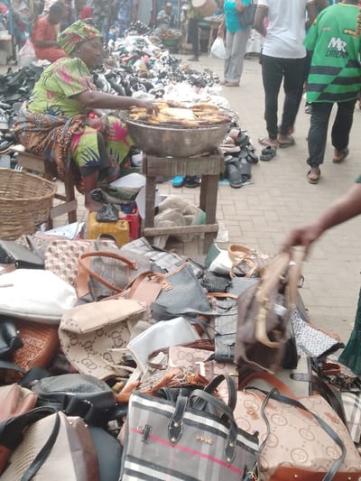 An open-air market scene shows a woman in traditional clothing cooking food on a grill. In the foreground, a large pile of handbags in various styles and patterns are displayed for sale. Additional merchandise, mainly shoes, spreads out in the background, blurring into the crowded marketplace. Shoppers move through the stalls, creating a bustling and active atmosphere.