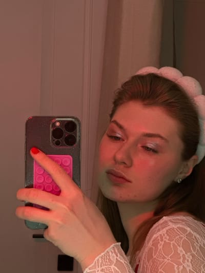 A woman poses in a selfie, wearing a lacy white top and a fluffy white headband. She looks downward with a slightly melancholic expression. Her hand holds a smartphone, which is reflecting the overhead lighting. The phone has a sparkling grey case and a pink, round-bubble grip on the back. The lighting creates a red-toned hue throughout the image.