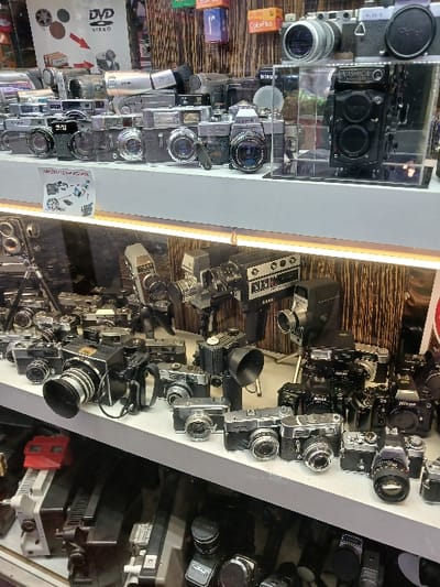 The image depicts a shopfront display in Istanbul, Türkiye, at the coordinates 41.022 latitude and 28.976 longitude. The display is located inside a shop, behind a glass window, and features a large collection of vintage cameras and other photographic equipment. The cameras are arranged on two shelves, one above the other, and are primarily silver and black in color. Some of the cameras are on stands, while others are stacked on top of each other. Several of the cameras have lenses pointed directly at the viewer, creating a sense of anticipation. The display also includes several other objects, such as a DVD video sign, and a variety of smaller items, including what appears to be an instruction manual for a camera. The overall impression is of a well-organized and carefully curated collection of vintage photography equipment. There are no people visible in the image.