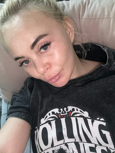 A blonde woman is lying down indoors. She is positioned on her side with her head resting on a pillow or cushion. She is wearing a dark-colored t-shirt with a white graphic print. Her eyes are open and she appears to be looking directly at the camera. The environment appears to be a relaxed, domestic setting, with a bed or couch visible in the background. The city Высокий, Russia is not visible in the image.