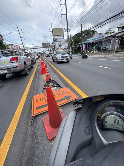 The image depicts a street scene in Wichit, Thailand, on a cloudy day. The viewpoint is from a scooter, looking forward and slightly to the right. Traffic cones and an orange directional sign are placed on the side of the road, indicating construction or a traffic diversion. Several vehicles, including cars and trucks, are visible in the traffic queue. On the right side of the road, there are shops and buildings with awnings. Two people are seen riding motorcycles in the distance. The scooter's dashboard shows a time of 6:52 and a speed of 0 km/h. The overall environment appears to be a moderately busy urban or suburban area.