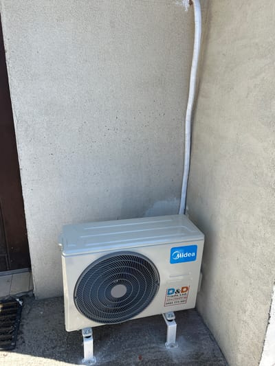 An outdoor unit of a Midea air conditioning system is mounted on white metal brackets on a concrete surface, positioned against a textured, light-colored exterior wall. To the left of the unit, a dark brown door frame is visible. A white insulated pipe runs vertically along the wall, originating from the top of the unit and disappearing upwards. A small sticker on the unit displays the "Midea" logo in blue and a company sticker with "Trade Ltd." and a phone number. The ground is concrete, with a black doormat visible near the door. The environment appears to be an outdoor space in Busmantsi, Bulgaria, likely a residential or commercial property.