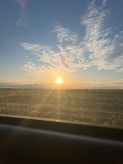 The image captures a serene sunset over a vast agricultural field, likely on the outskirts of Slobozia, Moldova. The sky is a brilliant canvas of blues and oranges, with wispy white clouds stretching across the horizon. The sun, a radiant orb, is positioned slightly to the left of the center, casting long, golden rays that illuminate the landscape. The field below is a tapestry of green and brown hues, with hints of vegetation and possibly distant trees or structures forming a faint silhouette against the setting sun. The foreground is dominated by a dark, blurry object, possibly a window frame or a car interior, suggesting the photograph was taken from a moving vehicle. The overall impression is one of tranquility and the natural beauty of a rural landscape at dusk.