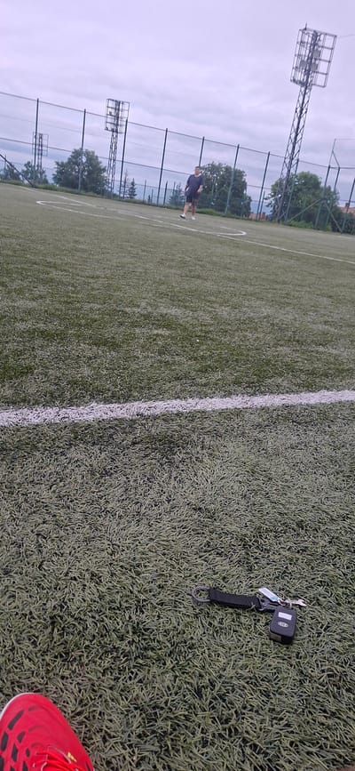 The image, taken in Lyaskovets, Bulgaria, displays an artificial grass sports field, likely a football pitch, under an overcast sky. The perspective is low, looking across the field from near the ground.

In the immediate foreground, on the bottom right, a person's foot clad in a vibrant red athletic shoe with black and white accents is visible. To the left of the foot and slightly further into the scene, a set of car keys lies on the green artificial turf. The keys include a modern black car remote fob, which appears to be for a Kia vehicle (the logo is faintly visible), attached to a keyring with a metallic key and a black strap keychain featuring a large silver ring.

A prominent white line, typical of sports field markings, runs diagonally across the mid-ground, extending from the bottom center towards the upper left. Further into the distance, on the left side of the field, a lone individual stands on the grass, wearing a dark t-shirt, shorts, and light-colored footwear, seemingly engaged in or observing an activity.

The background features tall, metal floodlight towers, indicating the field is equipped for night use, with one prominent in the upper left. A chain-link fence separates the field from a line of trees. The sky is a uniform, light grey, characteristic of an overcast day. The scene suggests a casual moment during or after a sports activity or training session on an outdoor pitch.
