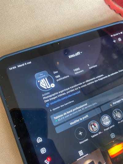 The image shows a close-up of a tablet screen displaying an Instagram profile. The profile belongs to "zou.off" and features the username "Zou," with 198 publications and 1082 followers. The bio describes the account as an "Artiste," focusing on intimate images worn on the body like fragments of memory, stemming from analog photography transformed into unique clothing. The tablet's interface shows options to add banners, view the professional dashboard, modify the profile, and share it. The battery level is at 54%. The tablet is lying on a beige surface. The date and time displayed on the screen are 14:54, Tuesday, May 5. There is no evidence to suggest it is Villeurbanne, France, so I will omit it.