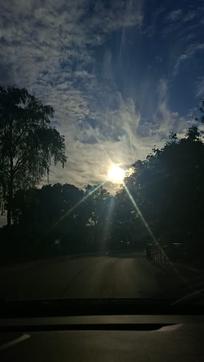 The image shows a view of the sky and trees from inside a vehicle, possibly on a road. The sun is visible, partially obscured by clouds and trees, casting light rays. The sky is a deep blue with scattered white clouds that have a wispy, feathery appearance. Silhouetted trees line the sides of what appears to be a road leading into the distance. The foreground shows the dashboard of the vehicle. There are no people or specific landmarks identifiable that would pinpoint the exact location within Smalyavichy, Belarus. The overall scene suggests a natural setting at daytime, possibly late afternoon or early evening due to the sun's position and the contrast between light and shadow.