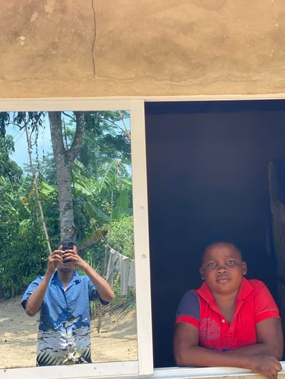 The image depicts a scene observed through and around a window in Ikot Ete Ekpo, Nigeria. The upper portion shows a light brown concrete wall with a prominent vertical crack and minor surface imperfections. Below the wall is a white-framed window, divided into two panes.
The left pane is reflective, mirroring an outdoor environment. Reflected in this pane is an individual, likely an adult male, holding a black smartphone vertically with both hands, appearing to be taking the photograph. He is wearing a blue short-sleeved collared shirt with a patterned lower section. The reflected outdoor scene includes a large tree with light-colored bark, dense green foliage consistent with banana plants, and patches of sandy ground under bright daylight. White or light-colored laundry is visible hanging on a line or bush in the reflected background.
The right pane of the window opens to a dark interior. From this interior, a young male looks directly towards the viewer, resting his forearms on the white windowsill. He is wearing a red short-sleeved polo shirt with dark blue trim on the sleeves. His expression is neutral.