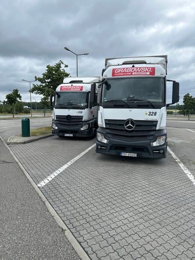 Two white Mercedes-Benz Actros semi-trailer trucks are parked side-by-side on a grey paver stone surface in a parking lot. The truck on the left has the number 338 displayed, and the truck on the right has the number 328. Both trucks have a red logo with white text that reads "GRABOWSKI FURNITURE LOGISTIC". The license plate on the truck to the left reads "PNT 66323", and the license plate on the truck to the right reads "PNT 65643". A green trash bin is visible to the left of the trucks. In the background, there are trees and streetlights. The sky is overcast. The image was taken in the city of Schipkau, Germany, on July 18, 2023.
