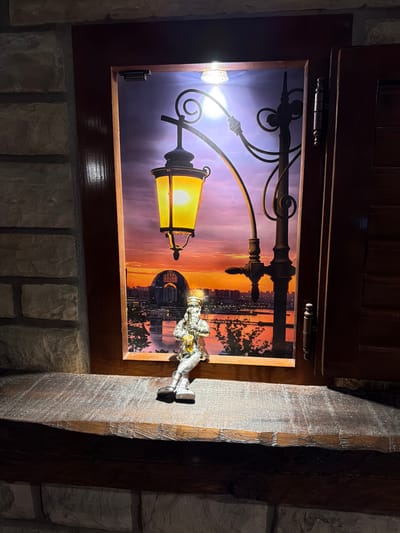 A small, silver figurine of a man playing a saxophone sits on a rough-hewn wooden ledge. The figurine is positioned in the foreground, facing left, with its legs dangling over the edge of the ledge. Behind the figurine, a framed picture depicts a city skyline at sunset, with a large, illuminated streetlamp in the foreground. The picture is housed within a wooden frame, which is slightly ajar, revealing a dark interior on the right. The ledge is set against a stone wall, with another section of stone visible below the ledge. The overall scene suggests an interior decorative display. The image does not provide any information that would indicate a location in Kumbor, Montenegro.