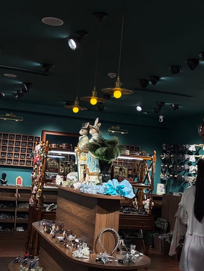 The image depicts the interior of a retail store specializing in hair accessories and decorative items. The store has dark teal walls and a dark ceiling with recessed lighting and several hanging pendant lights. The overall ambiance is somewhat dim, with spotlights highlighting displays of merchandise.

Prominently featured are two large mannequin-like figures of rabbits dressed in elaborate clothing, one wearing a top hat and the other a bonnet. These figures are positioned near the center of the image behind a wooden display counter. The counter itself is filled with various hair clips, headbands, and other small accessories, some of which are illuminated by a light strip beneath a shelf.

To the left of the rabbits, a wooden display unit with numerous small drawers or compartments holds an array of items, possibly earrings or hairpins. Further to the left, shelves are stocked with more merchandise.

On the right side of the image, a woman with long dark hair is partially visible, facing away from the camera. She appears to be a store employee or customer and is wearing a light-colored top and a light purple skirt. Behind her, a wall is lined with racks displaying numerous black and patterned hair accessories, such as bows and headbands.

The store appears to be well-stocked, with a variety of display shelves, counters, and hanging racks. Baskets are also visible, used for storage or display. The scene suggests a boutique setting, possibly aimed at a younger demographic or those interested in whimsical and decorative items. The location is identified as Heping District, China.