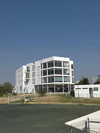 The image depicts a modern, multi-story white building under a clear blue sky in La Venta, Mexico. The building features extensive glass facades, particularly on its right side, with large, dark-paned rectangular windows arranged in a grid pattern across multiple floors. The left side exhibits white walls with smaller, irregularly spaced windows and two visible balconies with white railings. Dark circular markings are present on some white wall sections. In the foreground, a green artificial turf sports field occupies the lower left, complete with a white soccer goal and net. A white banner or tarp is visible behind the goal. The field is bordered by a dry, brown grassy area and a dark-colored fence. To the right of the sports field, closer to the main building, is a smaller, white rectangular structure with green signage and an entrance. Sparse trees and shrubs are scattered throughout the scene, primarily in the midground and background. No individuals are visible. In the immediate foreground, a partial white triangular structure and a metal fence are present on the lower right, indicating the vantage point.