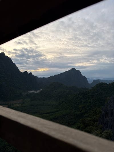 A wide, low-angle landscape photograph captures a view of lush green mountains and a cloudy sky through an overhead wooden railing. The railing, in the foreground, is a dark brown, blurred object that occupies the top and bottom edges of the frame, creating a tunnel-like effect.

The sky is filled with a layer of textured, cumulus clouds, ranging in color from white to light gray. Toward the horizon, a soft glow of yellow and orange suggests either sunrise or sunset.

The mountainous terrain is densely covered with dark green vegetation, with some areas appearing almost black due to shadow. A few wisps of white mist are visible in a valley between the mountains. The prominent peaks are jagged and steep, with some rocky outcrops visible on the right side of the frame. The scene is serene and natural, evoking a sense of remote wilderness.

The photograph is likely taken from an elevated vantage point, possibly a balcony or overlook, in or near Naka, Laos, a region known for its dramatic karst landscapes.
