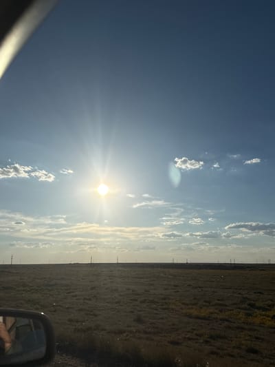 The image captures a vast, flat, arid landscape under a bright, sunny sky with scattered clouds. In the foreground, a dark car side mirror is visible on the left, reflecting a blurred image of a person's hand and arm. Beyond the mirror, the terrain is covered in dry, sparse vegetation, with some patches of yellow flora. Several tall, thin poles, likely power line supports, are spaced across the horizon, extending into the distance. The sky is a clear blue, with the sun prominently positioned in the upper left quadrant, emitting strong rays of light and causing a lens flare. The overall scene suggests an open, rural environment, possibly on the outskirts of a settlement, with the characteristic flat topography of the region. The location is identified as Töretam, Kazakhstan.