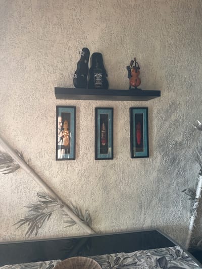 The image shows a textured, light-colored wall in what appears to be an indoor setting. A black shelf is mounted on the wall, displaying several items. On the left side of the shelf are two black objects that resemble a violin case and a bottle of Jack Daniel's whiskey. To the right of these is a small figurine of a person playing a cello. Below the shelf, three framed pictures are hung on the wall. The frames are black with a teal matting. The leftmost picture contains a photograph of a man holding a wooden object. The middle and rightmost pictures display what appear to be tribal masks, one red and one with dark and light brown colors. In the lower left and right portions of the image, parts of what look like decorative bamboo stalks with leaves are visible, extending from the bottom edge and partially onto the wall. The bottom edge of the image also reveals a dark surface, possibly a piece of furniture, with a patterned fabric. The overall ambiance suggests a decorated interior space. There is no discernible information in the image to indicate a specific location in Brca, Montenegro.