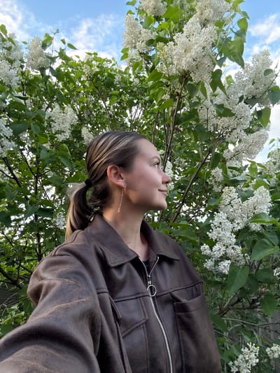 A low-angle shot captures a young woman looking upward towards a flowering tree, likely a lilac, with white blossoms and green leaves. The sky is visible through the foliage, displaying a pale blue color with some clouds. The woman is fair-skinned, and wears a brown jacket with a zipper. Her hair is pulled back into a low ponytail, and a light-colored streak is visible in her hair. She wears a dangling earring. The location is not evident from the image.