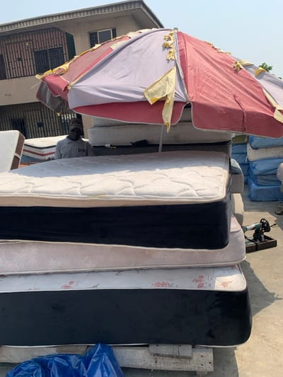 The scene depicts an outdoor commercial display in Alaiyabiagba, Nigeria, under bright direct sunlight. In the foreground and midground, a vertical stack of four mattresses dominates the frame. The uppermost mattress features a white quilted top and black side panels with white piping. Below it are two mattresses exhibiting light-colored, possibly beige or pink, floral-patterned fabric. The bottom mattress, resting on visible wooden pallets, also has a white quilted top with black side panels and white piping.
Providing partial shade over the mattress stack is a large, weathered market umbrella. Its fabric is faded red and light purple/grey, with numerous torn yellow strips of material.
Behind the umbrella and the central mattress stack, a male individual is partially visible. He wears a light-colored short-sleeved collared shirt with a dark logo (partially obscured, possibly "JUVAH") and a dark beanie hat with visible text (partially obscured, possibly "BOSS"). He appears to be looking forward.
To the right of the main mattress stack, a small, dark mechanical device, resembling a hand-cranked sewing or upholstery tool, rests on a black tray on the ground. In the background, a multi-story building with a light-colored facade and dark-framed windows with security bars is visible. Further to the right in the background, additional stacks of mattresses, some wrapped in blue plastic, are present. A crumpled blue tarp or plastic sheet lies on the ground in the lower-left foreground. The ground surface is light-colored, consistent with concrete or compacted earth.