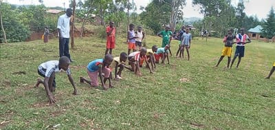 The scene depicts a group of children participating in a physical activity outdoors. The children are mostly in their teens and appear to be participating in a race. One child is kneeling on the ground and starting a race, while the rest of the children watch. There are two other children in the background, standing and looking at the children participating. There is one adult male standing on the edge of the frame. The scene is set in a grassy, open area, and there is vegetation in the background. The location is a grassy area in Bungoma, Kenya, near the coordinates 0.553 latitude and 34.773 longitude.