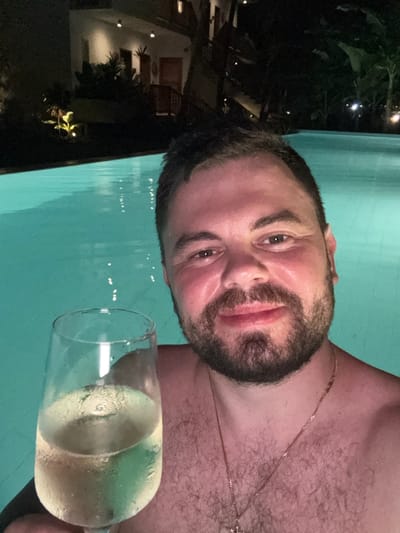 A light-skinned man with a dark beard and short dark hair poses for a selfie in a pool, a glass of white wine in his hand. He wears a delicate gold necklace. The pool water is light blue. In the background, a dark scene shows plants and what appears to be a building with lit windows. The image is slightly rotated.