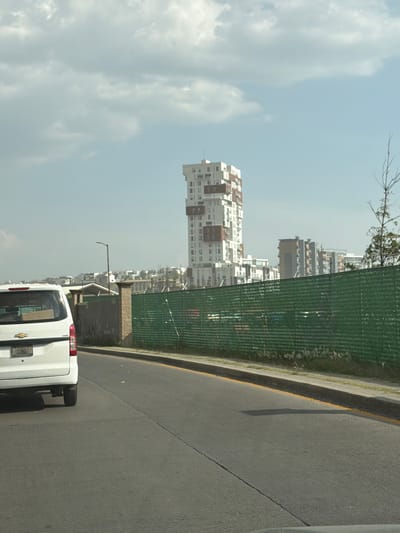 A slightly hazy, daytime street-level shot, rotated 90 degrees counter-clockwise, depicts an urban scene in Santa Clara Ocoyucan, Mexico. A white van is positioned on the right-hand side of the road. The road itself, composed of grey asphalt, has a visible yellow line marking the edge of the road. A green mesh fence runs alongside the road, separating it from a line of vegetation and the city beyond. Several multi-story buildings, characterized by white and brown exteriors, are visible in the background, indicating a dense urban environment. A light pole and a concrete utility structure are visible next to the fence. A cloudy sky is present on the left.