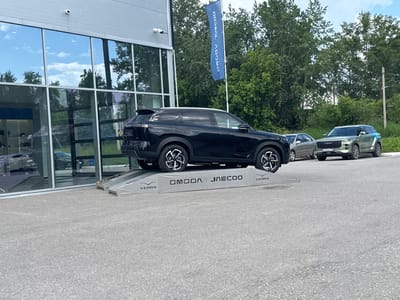 In the city of Berezniki, Russia, a black OMODA JAECOO SUV is parked on a grey ramp in front of a modern building with large glass windows. The ramp has "VERRA" and "OMODA JAECOO" branding. To the right of the black SUV, a lighter colored SUV with the same branding is parked on the asphalt. In the background, trees and a blue flag with white text are visible. The scene appears to be in a car dealership or service area.