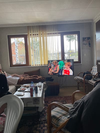 The scene depicts an interior, likely a living room, in Rupkite, Bulgaria, where two individuals are reclining on a sofa with their legs stretched upwards, their feet visible near a television screen mounted on the wall, which is showing a music performance. A table in the center of the room is laden with various items, including bottles of water, cans, glasses, plates, and a book. Chairs are scattered around the room, some with patterned cushions. Two windows with closed blinds let in natural light, revealing a view of trees outside, indicative of an early spring season. Above one window is a decorative picture. The room's aesthetic is dated, with a combination of patterned wallpapers and decorative wall tiles.