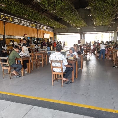 The scene is an outdoor restaurant in Kecamatan Ngaliyan, Indonesia, located at latitude -7.002 and longitude 110.342. It is partially covered with a leafy canopy. Several tables are set up with people sitting and eating. Many people are standing and talking near the service counter.  There is a large yellow sign with the phrase "If you stay hungry, cause hungry is rest". The floor is tiled, and there are yellow lines on the floor. Several people are standing around and others are seated in the shade, and most of the people are facing each other, likely interacting.  Some people are wearing hats, shirts, and trousers while others are wearing traditional clothing. There are several wooden tables and chairs set up.  A few people are carrying small bags.  The weather appears to be clear, and the light is bright.