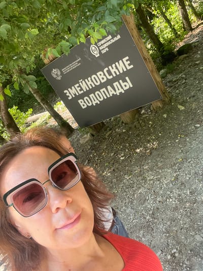A medium shot shows a woman in the foreground of a wooded area, looking up and taking a selfie. She is wearing sunglasses and a red top. Behind her, a sign in Russian reads "Zmeikovsky Waterfalls" and also displays the logo of "Sochi National Park." A second person is partially visible behind the woman, appearing to be leaning on a tree. The background consists of trees, foliage, and a gravelly path.