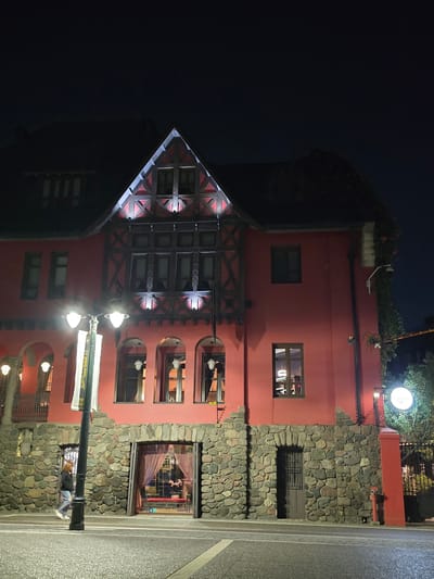 The image shows a distinctive building with a European-influenced architectural style, particularly with a prominent triangular roof, half-timbered facade elements, and decorative detailing. The building is primarily red in color, with stone accents along the base and sides. Several windows are visible, some illuminated from within, suggesting activity inside. A street light illuminates the building's facade. A section of the street is visible in the foreground. The building's design suggests a restaurant or commercial establishment.
Based on architectural characteristics, the location could be identified as the Baviera Restaurant in Providencia, Chile. The building is notable for its Bavarian-inspired design and is located in the Providencia area of Santiago, Chile.