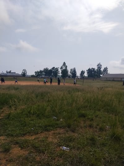The image depicts an outdoor scene with a group of young people engaged in what appears to be a sporting activity on a dirt field. Several individuals are running, some with athletic stances suggesting a race or practice. The setting includes a long, grassy field in the foreground and a dirt track or field in the mid-ground where the people are active. In the background, there are trees, buildings with corrugated roofs, and a few utility poles.
The sky is overcast with scattered clouds, indicating a possibly warm day, though the overall lighting suggests it is daytime. There are some scattered pieces of litter on the grass in the foreground, including a plastic water bottle. The overall mood appears to be casual and active, with the individuals seemingly focused on their activities. There is no discernible text visible in the image. The location context provided is Wumat, Nigeria.