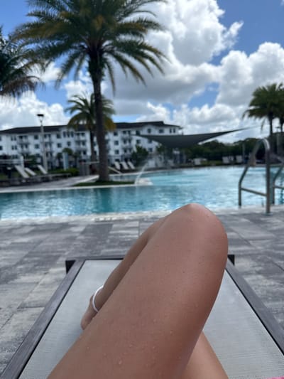 A close-up shot taken in what appears to be Naples, Florida, depicts a person relaxing on a lounge chair beside a pool. The foreground features a tanned leg with droplets of water, resting on a grey mesh lounge chair. An anklet is visible around the ankle.
The background reveals a swimming pool with a grey deck. Beyond the pool, palm trees sway in the breeze. A large, modern white building with a dark spire stands in the distance. The sky is blue with scattered white clouds.