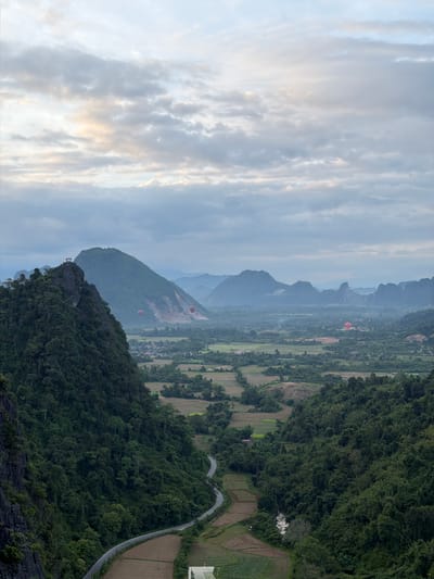 The image captures a stunning aerial view of a mountainous landscape in Naka, Laos. The scene is dominated by lush green forested hills and karst formations, indicative of a tropical or subtropical environment. In the distance, several hot air balloons are visible, adding a touch of adventure and a focal point to the vast scenery. One balloon, colored red, is prominently positioned over a valley, while others are smaller specks in the sky.

The terrain below reveals a patchwork of agricultural fields, some appearing to be rice paddies, with a winding road cutting through the dense vegetation. A small building with a white roof is nestled among the trees near the road. The perspective suggests the photograph was taken from a high vantage point, possibly another mountain peak or a hot air balloon.

The sky is partly cloudy, with soft, diffused light suggesting it is either early morning or late afternoon. The clouds are streaky and wispy, casting a gentle glow on the landscape. The overall atmosphere is serene and picturesque, evoking a sense of peace and wonder at the natural beauty of the location. There is no visible text in the image.