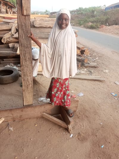 This full-shot photograph, taken in Ebiya, Nigeria, features a smiling young girl standing outdoors in what appears to be a timber yard or a similar setting where wood is stored. The girl, of African descent, stands on a wooden structure, holding onto a thick, dark wooden post to her left with her right hand. She is wearing a light-colored (possibly off-white or cream) hijab or head covering that drapes over her shoulders, and a vibrant red skirt adorned with a pattern of light green geometric shapes. She wears dark, open-toed sandals on her feet. Her face is turned towards the camera, displaying a wide, cheerful smile.
In the background, large quantities of rough-hewn wooden planks and logs are stacked, suggesting the primary activity of the area. To the immediate left, an old tire rests on the sandy ground. Partially visible behind the wooden post and the girl is another person, seen only from the back of their head (wearing a white cap) and their feet, near which a small broom is visible, indicating some activity like sweeping. To the right of the girl, a paved road or dirt track runs alongside more stacked logs and a sandy embankment leading up to dry, sparse vegetation and hills in the distance. The ground throughout the scene is sandy and appears somewhat cluttered with small pieces of litter and debris.
The lighting suggests it is daytime, possibly late morning or afternoon, with soft illumination and no harsh shadows, indicating clear, dry weather. The girl's modest attire aligns with cultural practices in the region. The image primarily captures a moment of cheerful interaction with the camera amidst an active, utilitarian environment. There is no visible text in the photograph.