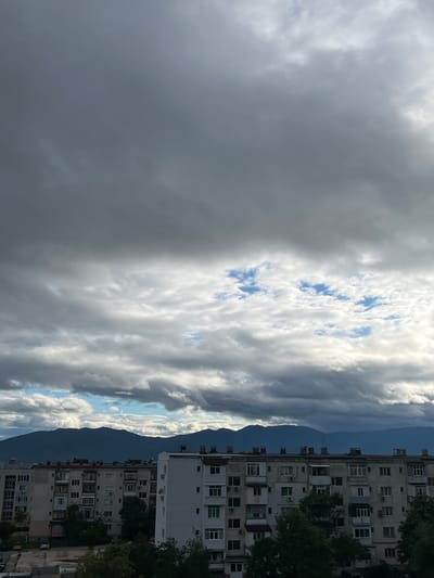 The image depicts a cloudy sky with a mountain range visible in the background, taken from a location in Sopot, Bulgaria, as implied by the presence of apartment buildings. The sky is divided between dark, ominous clouds on the left and lighter, patterned clouds with patches of blue on the right. The mountains appear dark and indistinct. The apartment buildings are white, multi-story structures with numerous windows and balconies. Some trees are seen alongside the building and in the foreground.