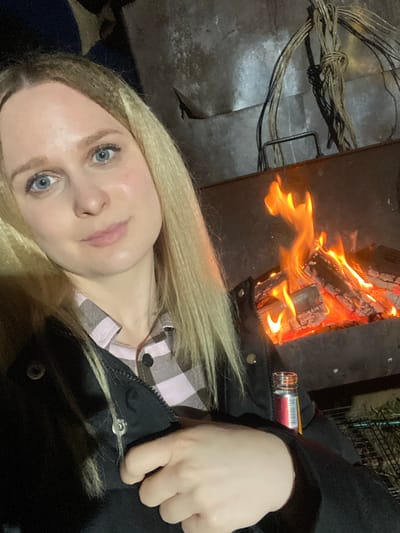 A self-portrait, taken at night, features a woman with long blonde hair and light blue eyes, looking directly at the camera with a neutral expression. She is partially visible from the chest up, wearing a black jacket over a pink and white checkered shirt. Her left hand rests near the collar of her jacket.

In the background, a warm, bright fire burns within a dark metal fire pit or fireplace. Several logs are engulfed in orange and yellow flames, illuminating the immediate surroundings. To the left of the fire pit, on a dark, possibly metallic surface, lies a tightly bundled coil of wires or cables.

A small, clear glass bottle containing a reddish liquid is visible near the fire, resting on the jacket or an adjacent surface. The bottle has a partially visible label with white and red elements, showing some illegible text and numbers.

The scene suggests an outdoor or semi-outdoor setting at night, likely in a cool environment, indicated by the use of a fire for warmth and light, and the subject's jacket. The dark background confirms it is nighttime. The location context of Kauchuk Stantsiya, Russia, implies a colder climate suitable for a fire. No strong emotions are discernible on the woman's face. No clearly readable text is present on the bottle's label.