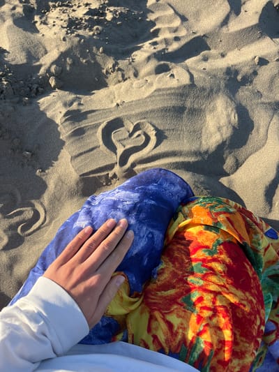 In the city of Pistula, Montenegro, a top-down, close-up shot captures a hand resting on a colorful fabric on a sandy beach. The sand, appearing light brown and textured with ripples and shadows, forms the background and foreground. A heart shape has been drawn into the sand, with the point facing towards the bottom right of the frame. The hand, belonging to a person wearing a white sweatshirt, is placed on top of two folded fabrics: one a vibrant blue with white swirls, and the other a multi-colored pattern of red, orange, yellow, and green. The scene suggests a peaceful and romantic moment on a sunny day at the beach.