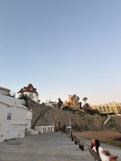 This image captures a view of the coastline in Cascais, Portugal, likely near the Praia da Rainha or a similar beach area. The scene is set during what appears to be late afternoon or early evening, with the sky a clear, pale blue, transitioning to a softer hue at the horizon.
In the foreground, a paved promenade runs along the edge of a sandy beach. Benches are arranged along the promenade, with a few individuals seated on them, enjoying the view. To the left, a white building with a blue tile plaque is visible, featuring windows and a slightly weathered facade. Further along the promenade, a row of small structures, possibly public restrooms or changing rooms, are lined up against a stone retaining wall.
Dominating the middle ground is a dramatic cliff face, constructed from rough-hewn stones. A set of stairs ascends this wall, leading to buildings perched at the top. These buildings include a white structure with a distinctive red-tiled roof, featuring multiple gables and dormer windows, and a tall, bright yellow building with arched windows and balconies, reminiscent of a hotel or large residence. Palm trees are scattered throughout this upper section, adding to the coastal ambiance. In the background, more yellow buildings with balconies can be seen, suggesting a developed coastal town.
Two people are the most prominent subjects in the foreground, seated on a bench facing away from the camera. A man with gray hair and sunglasses is wearing a maroon shirt, while a woman beside him has long dark hair and is wearing a white top and patterned shorts. They appear to be in conversation, looking towards the sea. Other figures are more distant and less distinct, scattered along the promenade and on the beach. The overall impression is one of a peaceful seaside setting, with a blend of natural landscape and architectural elements.