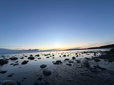 The image depicts a rocky shoreline at dusk in Haabneeme alevik, Estonia. The sky is a gradient of deep blue to light blue, with a faint orange and yellow glow on the horizon. Scattered across the dark, wet sand and shallow water are numerous dark, smooth rocks of varying sizes. The water reflects the colors of the sky. In the distance, a silhouetted landmass with trees and a small structure is visible. There are no people or animals in the frame. The overall scene is serene and natural.