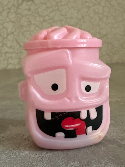 A pink toy with a carved brain on its head sits on a textured gray surface. The toy's face has two large white eye sockets with black pupils, a wide mouth with black interior, and several white teeth. A red tongue protrudes from the mouth. The background is a textured, light beige wall. The image was taken in Orsk, Russia.