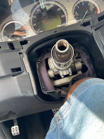 The image depicts the interior of a vehicle from the driver's perspective. The specific location in Alaiyabiagba, Nigeria, is not discernible from the image. The steering wheel is absent, exposing the silver-colored, threaded steering column shaft and associated mechanical components, surrounded by black trim and purple-grey insulation material. Above, the vehicle's instrument cluster is visible, showing a speedometer peaking at 160 units and a tachometer up to 7,000 RPM, beneath a clear protective cover. To the lower left, a metallic, perforated pedal is visible. In the lower right quadrant, a person's right leg, clad in blue denim, is present in the footwell area. The scene indicates the vehicle is in a state of disassembly or repair.