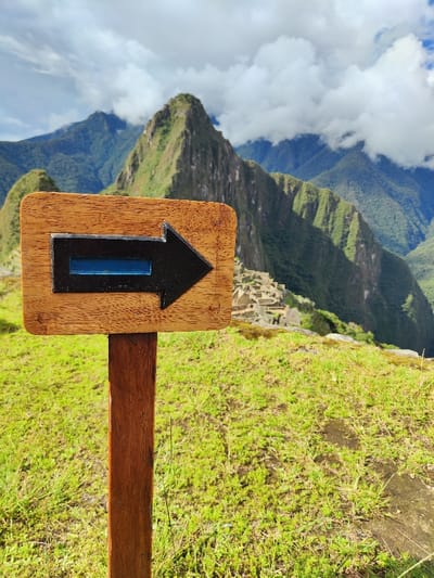 A wooden sign with a black arrow pointing right stands in a grassy field. The arrow contains a blue rectangle. Behind the sign, a mountainous landscape with green slopes and rocky peaks rises into a cloudy sky. In the background, hints of ancient structures and ruins are visible, suggesting a historical or archaeological site. The photograph captures a sense of adventure and exploration in a remote, scenic location.