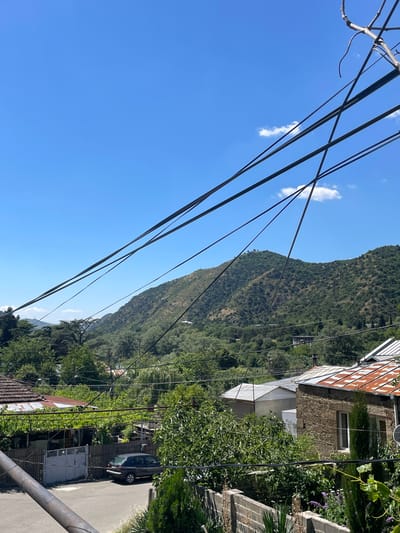 The image captures a bright, sunny day in Mtskheta, Georgia. The foreground is dominated by a jumble of electrical wires crisscrossing the frame, obscuring a clear view of the scene below. Beyond the wires, a residential area is visible, characterized by houses with tiled roofs, some of which appear weathered and aged. Lush green trees and vegetation are abundant, suggesting a warm climate and well-established gardens. A dark-colored car is parked on a paved area. In the background, rolling green hills, covered in dense foliage, rise up, forming a natural backdrop to the human settlement. The sky is a clear, vibrant blue, with only a few wispy clouds scattered across it. The overall impression is one of a quiet, somewhat rural, or suburban setting within the historic city of Mtskheta.
