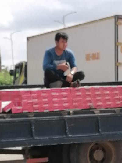 A blurry, medium-shot, eye-level photograph shows a person sitting cross-legged on a flatbed truck, facing right. The person, likely male, is wearing a blue long-sleeved shirt, dark pants, and dark shoes. A watch is visible on the person's left wrist. The person is holding an object in their right hand, possibly a cigarette or a lighter. The truck is loaded with rows of pink boxes, stacked neatly. In the background, a white truck trailer is visible, with some text in yellow that is unreadable due to the blur. The environment appears to be outdoors, with a cloudy sky. The presence of the flatbed truck, stacked goods, and the trailer suggests this scene might be related to transport or logistics, possibly in an industrial or commercial area. Given the prompt's constraint, the location is identified as Xuân Hương - Đà Lạt, Vietnam.