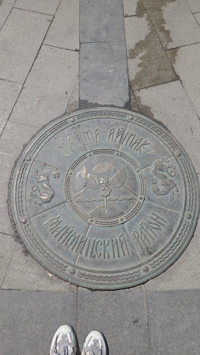 The image shows a circular bronze plaque embedded in a gray stone pavement, with the feet of a person wearing white sneakers visible at the bottom. The plaque features relief carvings and text in Cyrillic script. At the center of the plaque is a stylized emblem depicting a winged owl holding a key, with a sun and wavy lines below. Around this central emblem, there are four quadrant-like sections. Two sections contain carvings of feline creatures, possibly leopards or snow leopards, in a dynamic pose. The text inscribed on the outer ring of the plaque reads "МАЙМА АЙМАК" on the top half and "УЛАГАНСКИЙ РАЙОН" on the bottom half.

This plaque is located in Gorno-Altaysk, Russia, and represents the symbol of the Mayma Aimag and Ulagansky District.